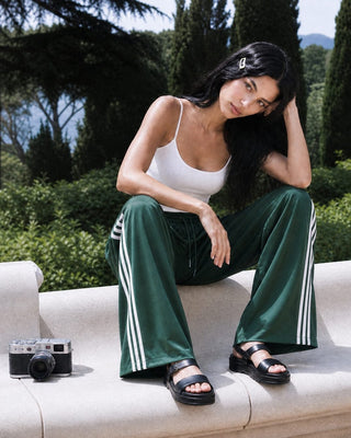 A woman sits on a light-colored stone bench outdoors, wearing dark green wide-leg track pants with white side stripes, a white tank top, and black sandals. Trees and greenery fill the background, and a small camera rests on the bench beside her.