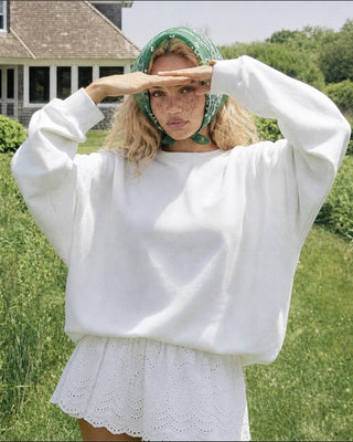 A woman stands outdoors in a grassy field wearing a loose white sweatshirt and a white skirt, with a green patterned headscarf. A house and trees are visible in the background.