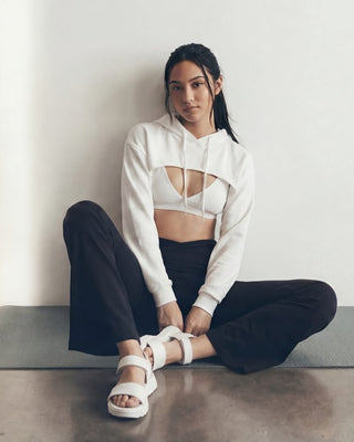 woman sitting on yoga mat in studio setting wearing cropped white hoodie, flare yoga pants and chunky white slides.